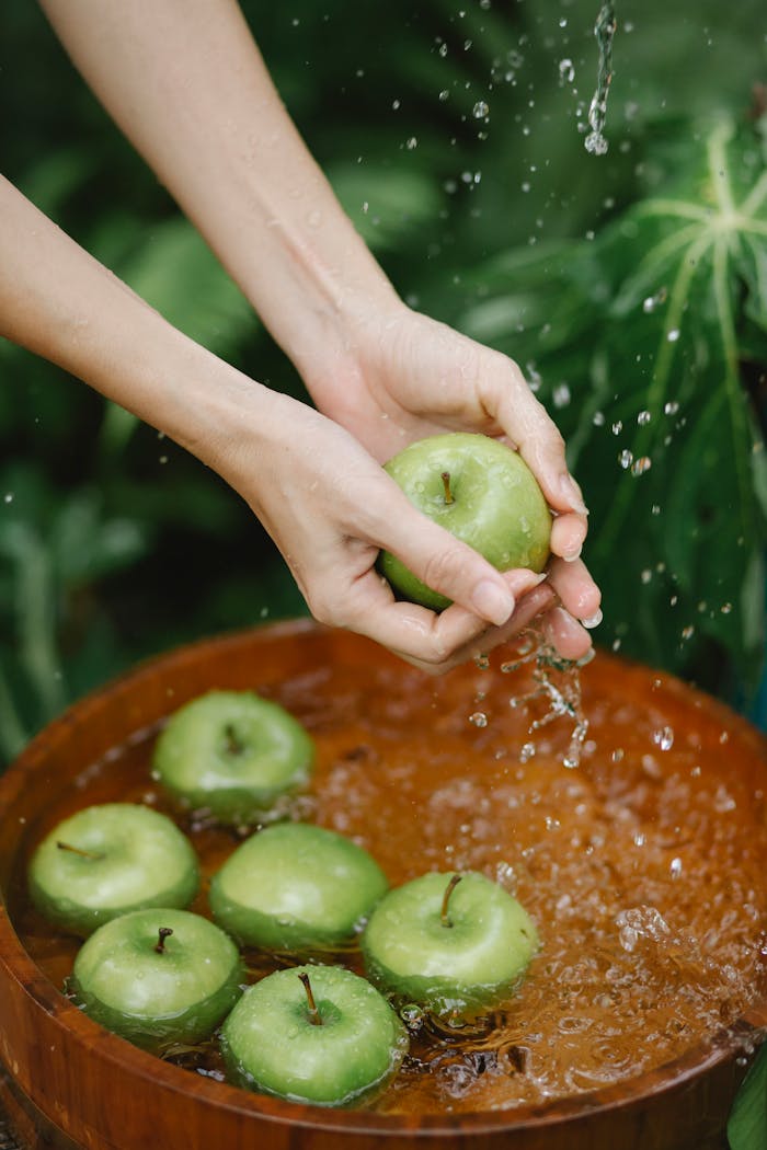 Hands wash fresh green apples in a wooden basin outdoors, surrounded by lush greenery.